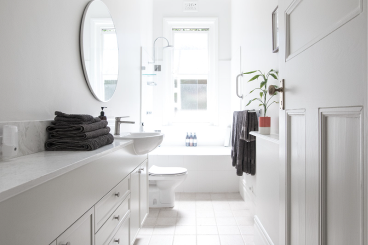 Bright white bathroom with a modern vanity, round mirror, bathtub, folded towels, and natural light from large windows.